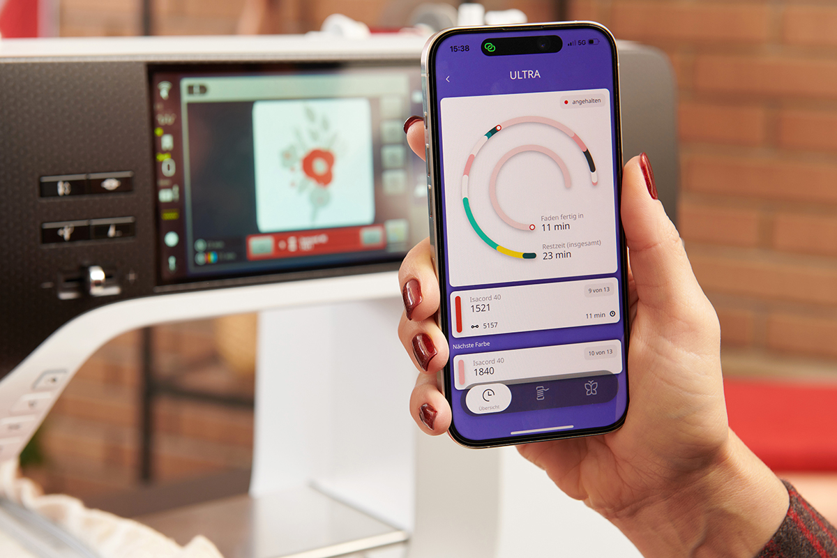 A hand holds up a smart phone in front of a sewing machine. The phone shows an app that illustrates the status of stitching on the machine beside it.