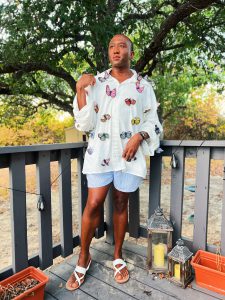 Black man wearing a handmade oversized white button-front linen shirt decorated with colorful embroidered butterflies, styled with blue striped shorts and white sandals outdoors.