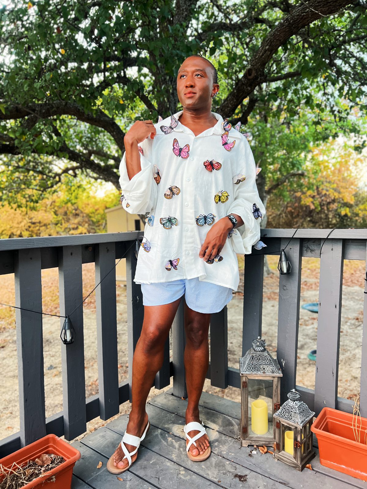 Black man wearing a handmade oversized white button-front linen shirt decorated with colorful embroidered butterflies, styled with blue striped shorts and white sandals outdoors.