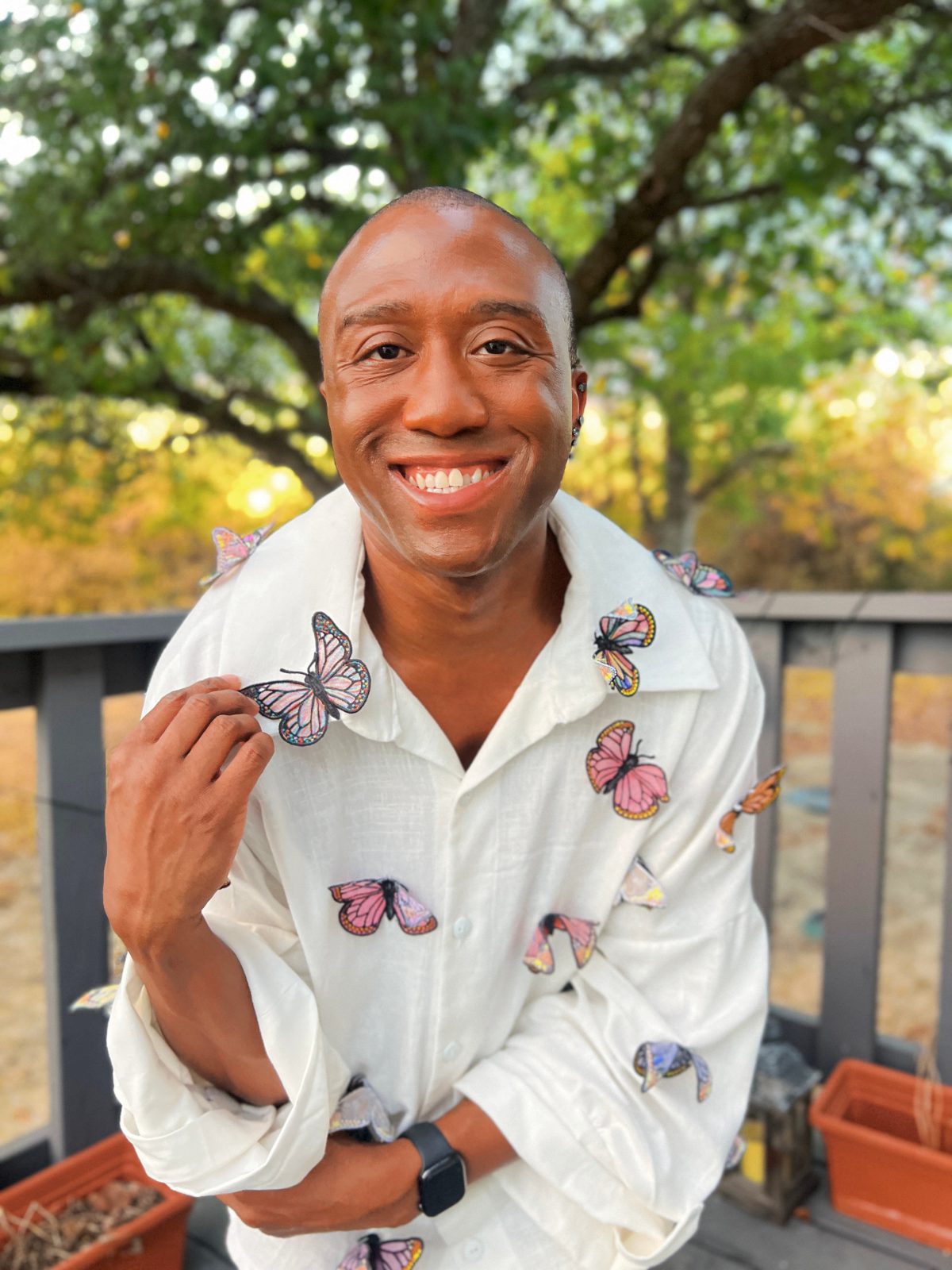 Black man wearing a handmade oversized white button-front linen shirt decorated with colorful embroidered butterflies, styled with blue striped shorts and white sandals outdoors.