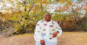 Smiling Black man modeling an oversized white linen button-front shirt covered in colorful three-dimensional embroidered butterflies against a backdrop of autumn trees.