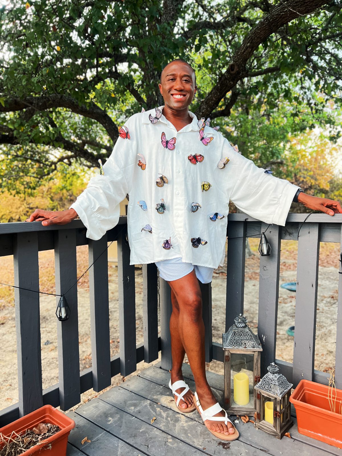 Smiling Black man wearing an oversized white button-front shirt embellished with colorful embroidered butterflies, paired with striped shorts and white sandals outdoors.
