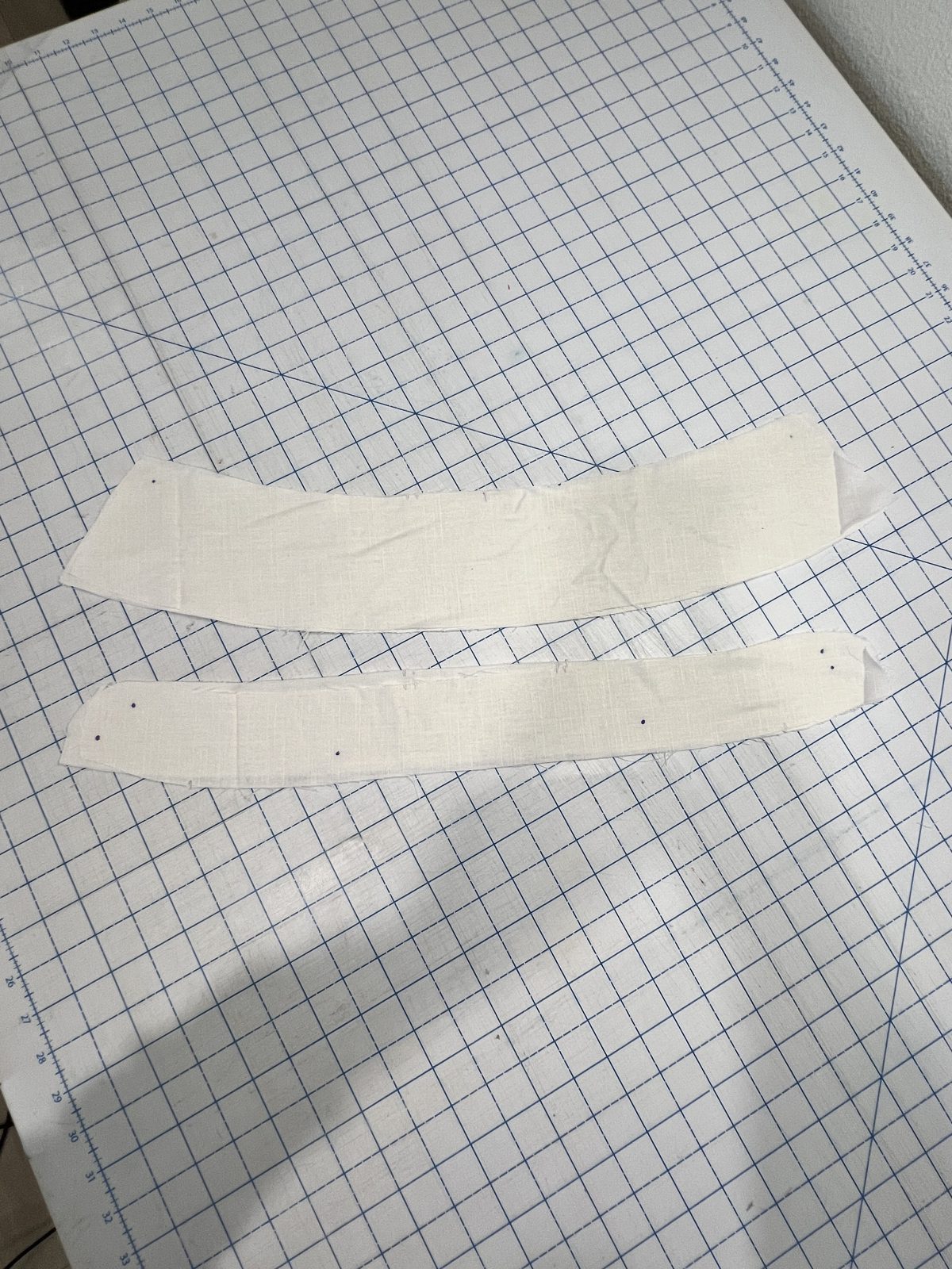 Two curved white linen collar pieces pinned and prepared for sewing on a gridded cutting mat.