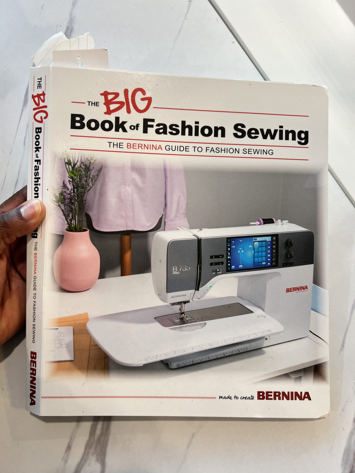 BERNINA’s The BIG Book of Fashion Sewing held over a sewing table, featuring a BERNINA sewing machine on the cover.