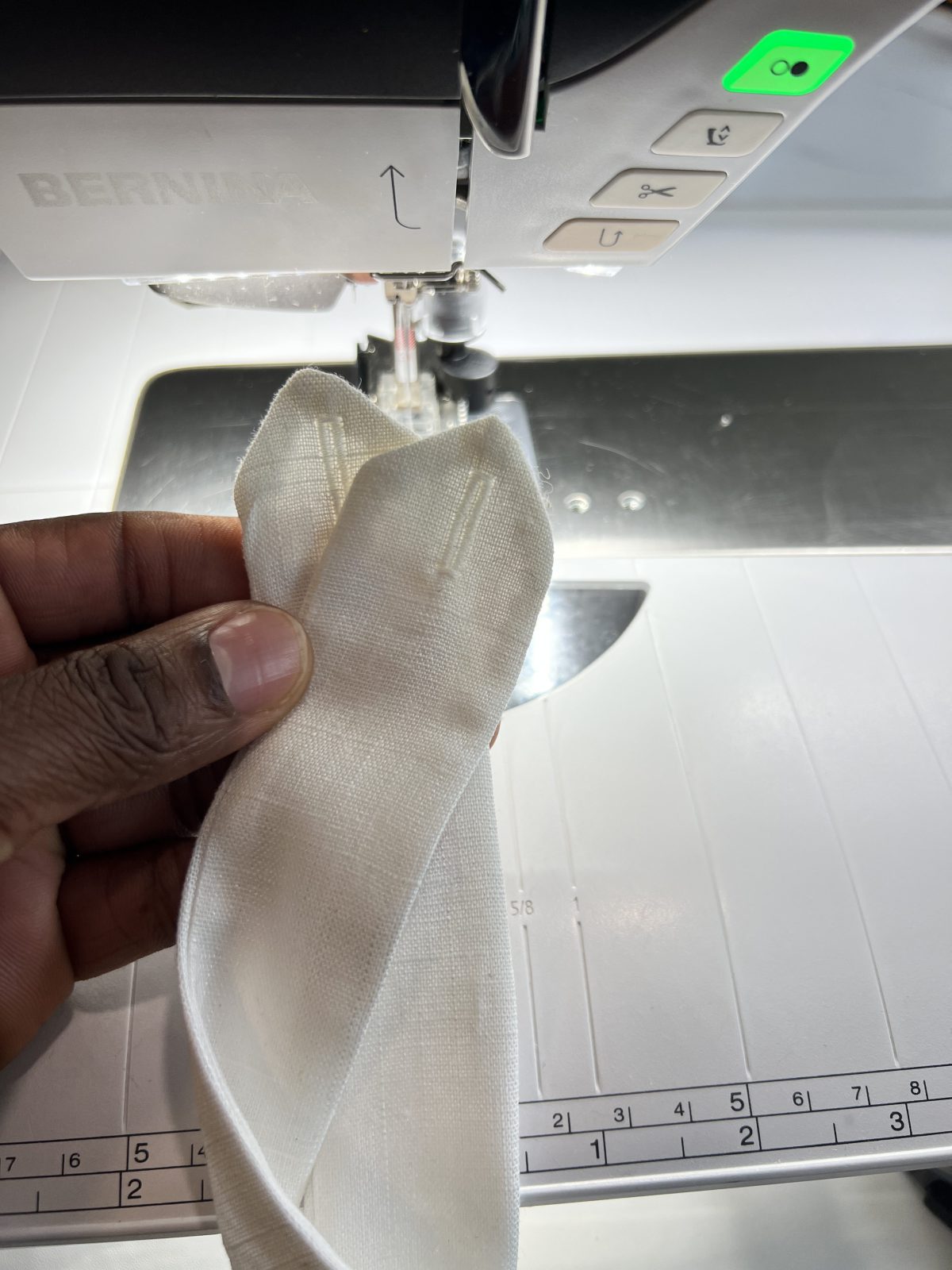 Close-up of two neatly stitched buttonholes on white linen fabric in front of a BERNINA sewing machine.