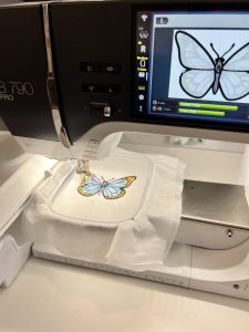 Blue and yellow butterfly embroidery design being stitched onto white fabric with a BERNINA B 790 PRO embroidery machine.
