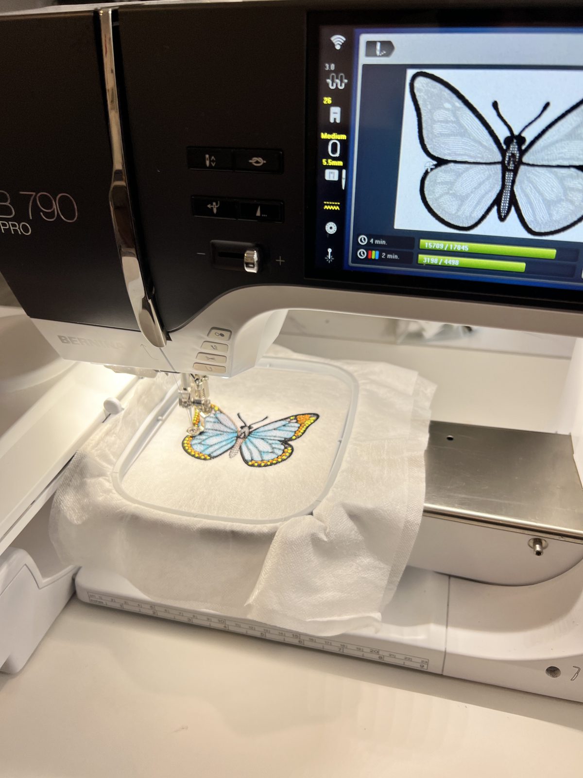 Blue and yellow butterfly embroidery design being stitched onto white fabric with a BERNINA B 790 PRO embroidery machine.