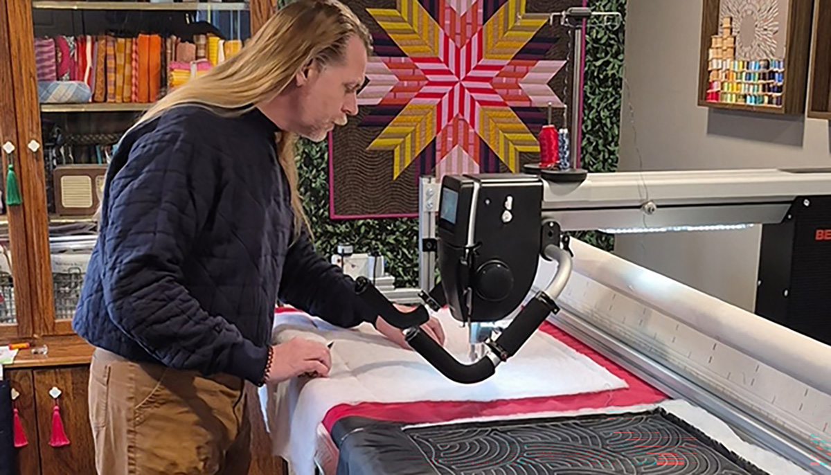 male standing at longarm inspecting a quilt